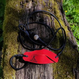 May include: A red leather pouch for a slingshot featuring a black logo with the text ONE STONE, attached to a black cord with a small light-colored bead. The cord is looped and tied, forming a functional projectile launching mechanism.
