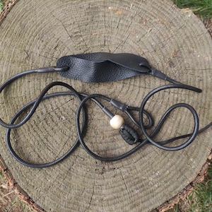 May include: A black leather slingshot featuring a textured leather pouch connected to two long black cords. One cord includes a smooth, light-colored wooden bead and a black knotted end piece, designed for manual tension and projectile launching.