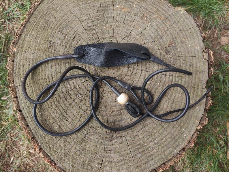 May include: A black leather slingshot featuring a textured leather pouch connected to two long black cords. One cord includes a smooth, light-colored wooden bead and a black knotted end piece, designed for manual tension and projectile launching.