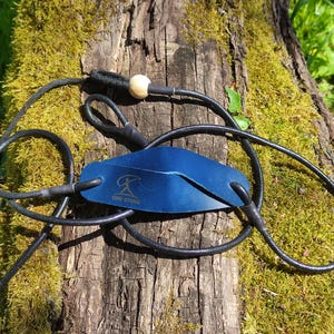 May include: A blue leather pouch for a sling with a gold logo that reads ONE STONE. The pouch is attached to black cords that form loops, with a small light-colored wooden bead secured at the top of the cord assembly for adjustment.