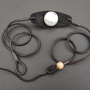 May include: A black leather pouch attached to two long black cords, featuring a white golf ball with the text INSIA printed on it. The cords are connected by a knot and a single light-colored wooden bead, forming loops at the ends of the cords.