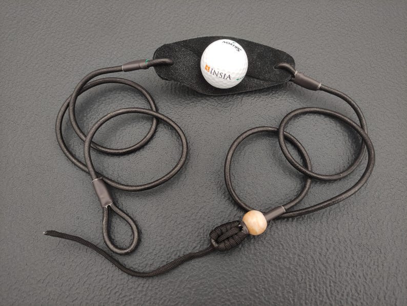 May include: A black leather pouch attached to two long black cords, featuring a white golf ball with the text INSIA printed on it. The cords are connected by a knot and a single light-colored wooden bead, forming loops at the ends of the cords.