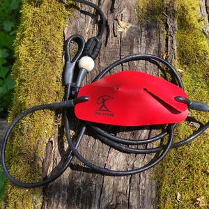 May include: A red leather pouch for a slingshot with a black cord and a white bead. The pouch features a black logo of a person throwing a stone with the text ONE STONE printed below it. The black cord is looped and attached to the red leather.