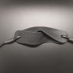 May include: A black leather camera strap featuring a textured, pebbled finish on the central pad. The strap is constructed with a rounded cord design that connects to the wider, contoured neck pad through reinforced, dark-colored attachment points.