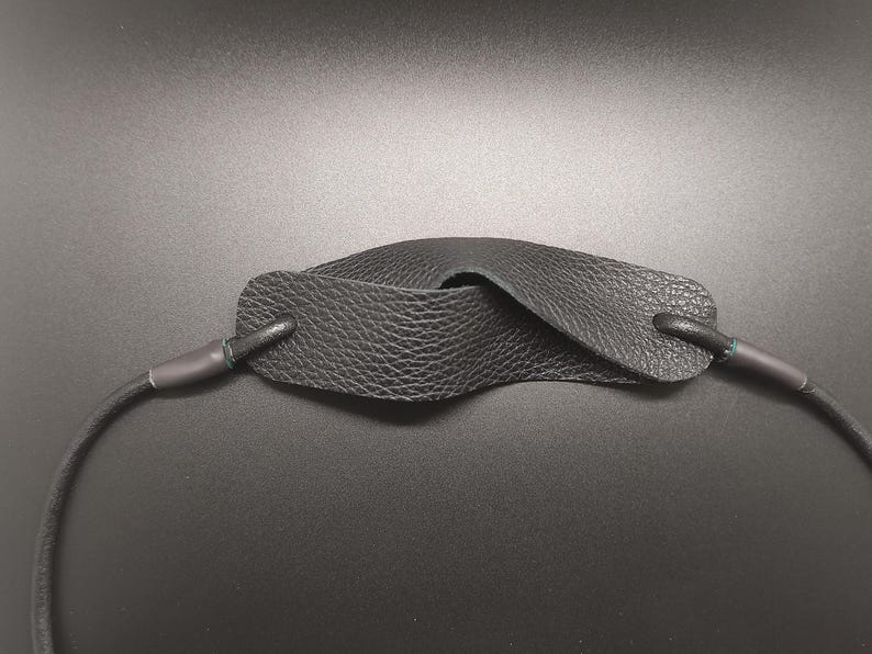 May include: A black leather camera strap featuring a textured, pebbled finish on the central pad. The strap is constructed with a rounded cord design that connects to the wider, contoured neck pad through reinforced, dark-colored attachment points.