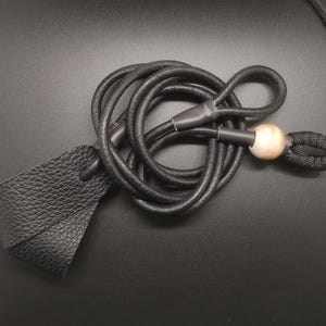 May include: A black textured leather loop attached to a long black cord. The cord features a smooth, round, light-colored bead and a black woven knot detail, with black plastic ferrules securing the connections between the leather and the cord.