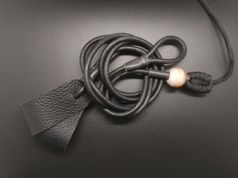 May include: A black textured leather loop attached to a long black cord. The cord features a smooth, round, light-colored bead and a black woven knot detail, with black plastic ferrules securing the connections between the leather and the cord.