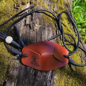 May include: A brown leather pouch for a slingshot featuring a black logo with the text ONE STONE. The pouch is attached to black elastic cords and a black cord loop with a single white bead. The leather is smooth with a dark gradient finish.