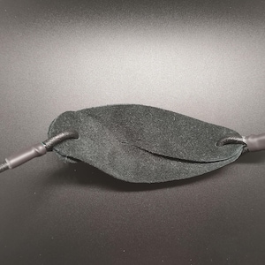 May include: A black leather pouch for a slingshot, featuring a textured surface and two holes on either side. The pouch is attached to two black elastic cords, each secured with a black heat-shrink tubing sleeve at the connection points.