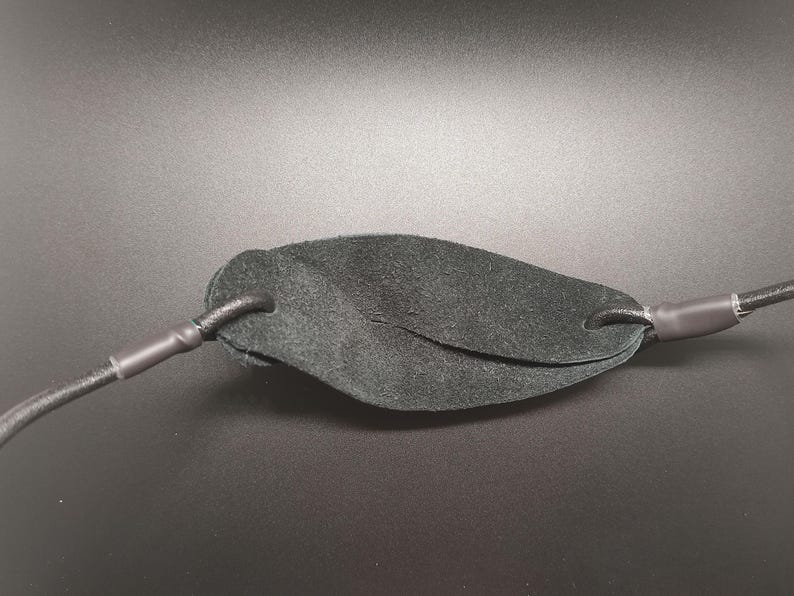 May include: A black leather pouch for a slingshot, featuring a textured surface and two holes on either side. The pouch is attached to two black elastic cords, each secured with a black heat-shrink tubing sleeve at the connection points.