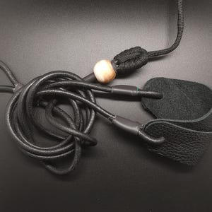 May include: A black leather cord with a textured surface, featuring a looped end and a black leather tab attached to the other side. A single round, cream and brown marbled bead is threaded onto the cord, secured by a black knotted paracord section.
