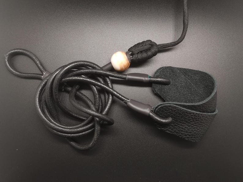 May include: A black leather cord with a textured surface, featuring a looped end and a black leather tab attached to the other side. A single round, cream and brown marbled bead is threaded onto the cord, secured by a black knotted paracord section.