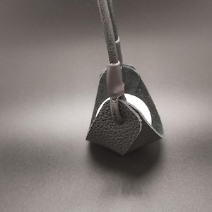 May include: A black textured leather sling holder featuring a rounded base and two vertical straps that meet at the top. A smooth white spherical object is cradled within the center of the black leather structure, which is held together by dark loops.