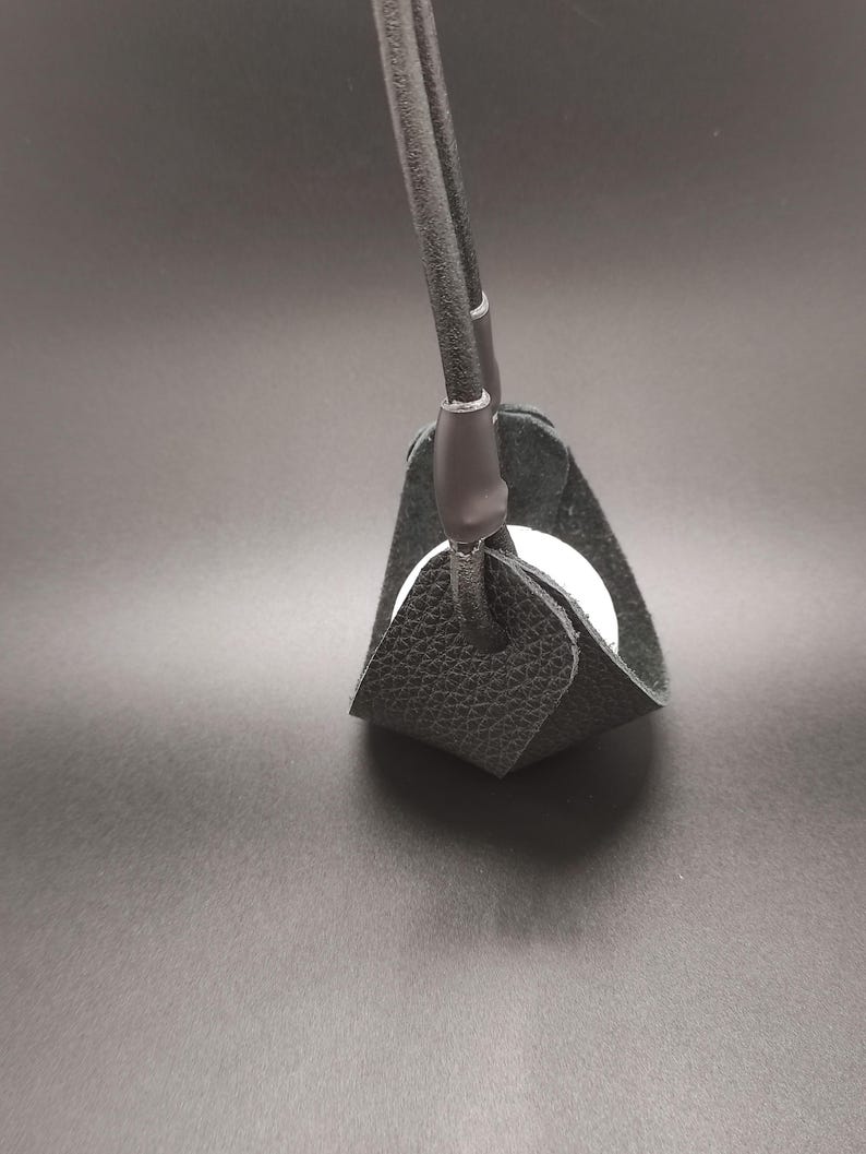 May include: A black textured leather sling holder featuring a rounded base and two vertical straps that meet at the top. A smooth white spherical object is cradled within the center of the black leather structure, which is held together by dark loops.