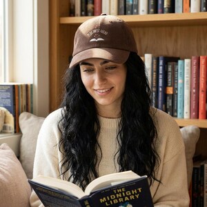 Puede incluir: Gorra de béisbol de pana marrón con el texto blanco bordado "Plot Twist Ahead". La gorra tiene una visera curva y un diseño clásico de seis paneles. La persona lleva un jersey color crema y está leyendo un libro.