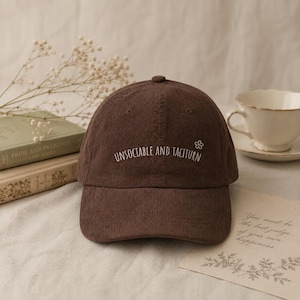 Puede incluir: Una gorra de béisbol de pana marrón oscuro con el texto blanco bordado "UNSOCIABLE AND TACTITURN" en el panel frontal, acompañado de un pequeño icono de flor blanca bordado situado a la derecha del texto.