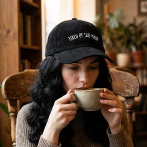May include: Black corduroy baseball cap with the text "TIRED OF THIS PSYOP" in white lettering. The cap is worn by a person drinking from a beige mug. The background includes a wooden bookshelf and a wooden chair.