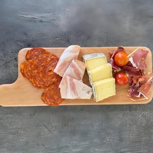 Può includere: Un tagliere in legno con un assortimento di cibo. Chorizo ​​a fette, prosciutto, formaggio e pomodori sono disposti sul tagliere. Il tagliere è marrone chiaro e ha un'estensione a forma di manico sul lato sinistro.