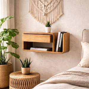 Puede incluir: Una mesita de noche flotante de madera clara, con un cajón y una estantería abierta. Está fijada a una pared de color neutro. Hay una pequeña planta en maceta y libros en la estantería. La habitación está decorada con plantas y un tapiz de pared.