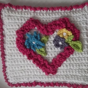 May include: A handmade, square crochet item with a white base and a vibrant pink heart-shaped design in the centre. The heart frames a cluster of colourful flowers, including blue, yellow, and purple. The edges are trimmed with a pink border.