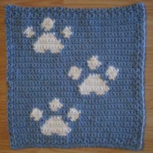 Op de afbeelding: Een vierkante, gehaakte blauwe washand met drie witte pootafdrukken. De washand is gemaakt van een getextureerd garen en de pootafdrukken zijn gelijkmatig over het oppervlak verdeeld. Het ontwerp is eenvoudig en strak.