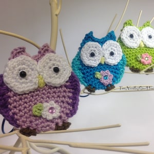 May include: Three crocheted owls with white eyes and beaks, each with a different colored body: purple, blue, and green. Each owl has a small flower with a rhinestone center attached to its chest. The owls are perched on a white branch.