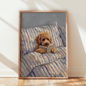 Op de afbeelding: Een ingelijste olieverfschilderij-print met een krulharige goudbruine puppy die rechtop op een bed met blauw-wit gestreept beddengoed zit. De puppy houdt een open boek vast met zijn voorpoten en kijkt geconcentreerd naar de pagina's.