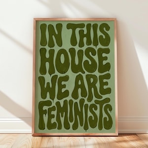 May include: A rectangular framed wall art print featuring a light sage green background with dark olive green text that reads IN THIS HOUSE WE ARE FEMINISTS. The lettering is rendered in a bold, wavy, and dripping liquid-style font.