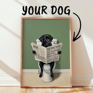 Puede incluir: Una lámina de arte enmarcada que muestra a un Labrador negro sentado en un inodoro blanco leyendo un periódico titulado THE DAILY WOOF con el titular LOCAL DOG DEMANDS BELLY RUBS, REFUSES TO MOVE. El fondo es una pared de color verde salvia.