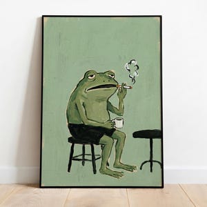 May include: Framed art print depicting a green frog smoking a cigarette and holding a coffee cup. The frog is seated on a black stool, wearing black shorts, with a second stool nearby. The background is a muted green.