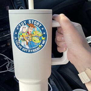 May include: A matte cream-colored insulated tumbler with a handle and a straw, featuring a circular Toy Story graphic with Woody, Buzz Lightyear, and two green aliens, along with the text Toy Story and You've Got A Friend In Me.