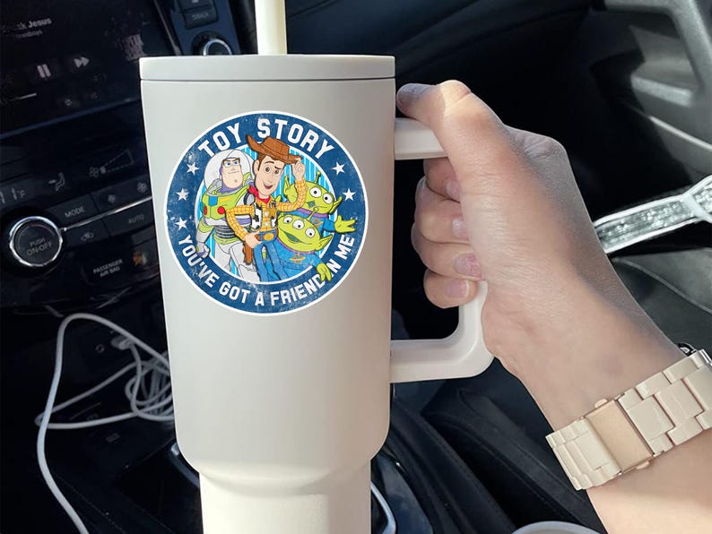 May include: A matte cream-colored insulated tumbler with a handle and a straw, featuring a circular Toy Story graphic with Woody, Buzz Lightyear, and two green aliens, along with the text Toy Story and You've Got A Friend In Me.