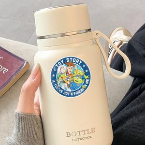 May include: A cream-colored insulated water bottle featuring a circular Toy Story sticker with Woody, Buzz Lightyear, and aliens. The text on the sticker reads TOY STORY and YOU'VE GOT A FRIEND IN ME. The bottle has a carry loop and BOTTLE HOT&COOL text.