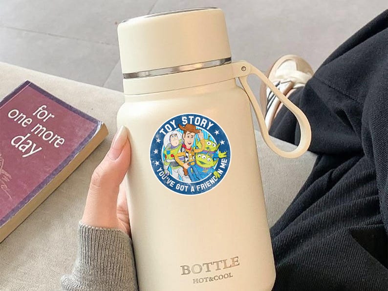 May include: A cream-colored insulated water bottle featuring a circular Toy Story sticker with Woody, Buzz Lightyear, and aliens. The text on the sticker reads TOY STORY and YOU'VE GOT A FRIEND IN ME. The bottle has a carry loop and BOTTLE HOT&COOL text.
