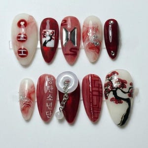 May include: A set of ten artificial nails in various designs. The nails feature a mix of red, white, and silver colors. Some nails have abstract patterns, while others display a tree, a logo, and Korean text.