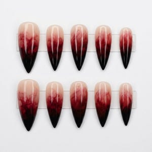 May include: A set of ten stiletto-shaped press-on nails featuring a gradient design that transitions from a nude base at the cuticle to a deep, smoky red and black ink-like marble effect toward the pointed tips, arranged in two rows of five.
