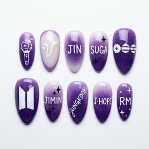 May include: A set of ten almond-shaped press-on nails featuring a purple gradient with glitter. Designs include white text reading V, JIN, SUGA, JIMIN, JUNGKOOK, J-HOPE, RM, a BTS logo, a lightstick icon, and star accents in black and white.