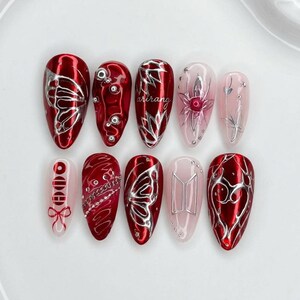 May include: A set of ten press-on nails featuring a mix of deep red metallic and translucent pink finishes. Designs include silver butterfly wings, raised silver beads, the word arirang, a floral motif, a red bow, a chain pattern, and a silver logo.