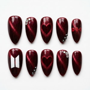 May include: A set of ten almond-shaped press-on nails in a deep burgundy cat-eye finish. The design features accents including clear rhinestones, a heart-shaped magnetic pattern, a white geometric logo, and small red circular embellishments.