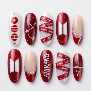 May include: A set of ten press-on nails featuring a red and white color palette with glitter accents. Designs include the BTS logo, the number 7, the word ARIRANG, star constellations, and ribbons printed with the names RM, JIN, SUGA, J-HOPE, JIMIN, V, and JK.