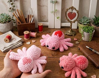 May include: Three handmade crochet spiders in varying shades of pink, each with a heart-shaped detail. One spider is held in open hands. The spiders are on a wooden surface with craft supplies and decorative elements.