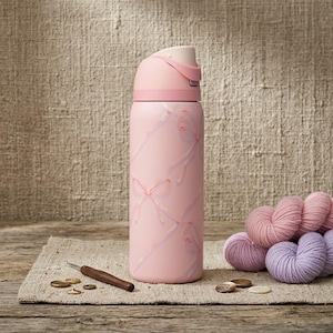 K&ouml;nnte beinhalten: Eine rosa isolierte Wasserflasche mit einem Schleifen- und Banddesign. Die Flasche hat einen hellrosa Deckel und einen passenden rosa K&ouml;rper. Ein H&auml;kelhaken, Garn und Kn&ouml;pfe befinden sich neben der Flasche.