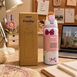 K&ouml;nnte beinhalten: Eine wei&szlig;e Owala FreeSip Wasserflasche mit rosa Schleifenmotiven und dem Text "Hi there, I'm leak proof, I'm insulated, And I have a built-in straw." Ein Karton mit dem Owala-Logo und dem Text "FreeSip" befindet sich neben der Flasche. Die Flasche fasst 350 ml.
