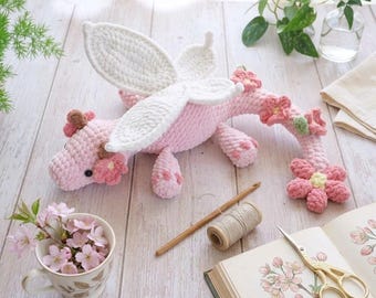 May include: A handmade pink crochet dragon with white wings and floral accents. The dragon is adorned with pink flowers and brown accents. A crochet hook, thread, and scissors are also visible.