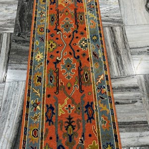 Könnte beinhalten: Ein rechteckiger Läuferteppich mit einem aufwendigen Muster. Der Teppich hat ein orangefarbenes Mittelfeld mit einem sich wiederholenden Design in Blau, Grün und Gelb. Der Rand ist blau mit floralen Motiven. Der Teppich ist wahrscheinlich aus Wolle.