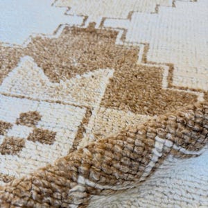 Könnte beinhalten: Ein rechteckiger Teppich mit einer dicken, hochflorigen Textur, einer cremefarbenen Basis und hellbraunen geometrischen Mustern. Das Design umfasst abgestufte Rautenformen und kleine quadratische Motive. Eine Ecke des Teppichs ist umgeschlagen und zeigt die Unterseite.