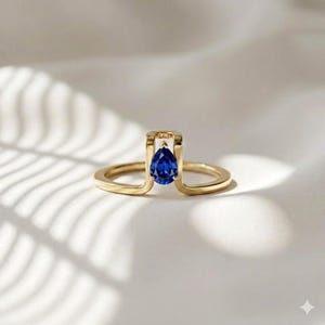 Könnte beinhalten: Ein goldener Ring mit einem tropfenförmigen, tiefblauen Edelstein. Das Design des Rings umfasst einen rechteckigen Rahmen um den Edelstein, mit einem zarten Band, das sich um den Finger legt. Der Ring ist vor einem weichen, weißen Hintergrund platziert.