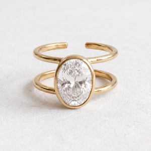 May include: A gold-toned open ring featuring a large, oval-cut clear gemstone set in the center. The band consists of two parallel gold-toned wires that connect to the central oval setting, creating a split-shank design with an open back.