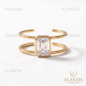 May include: A gold open-style ring featuring a central emerald-cut clear gemstone held in a bezel setting. The band consists of two parallel gold wires that connect to the central stone. The text ELANZIA appears multiple times in the image.