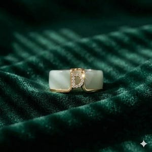 Könnte beinhalten: Ein goldener Ring mit einem hellgrünen Stein und einem diamantbesetzten Buchstaben "D" in der Mitte. Der Ring hat eine rechteckige Form mit abgerundeten Kanten, vor einem dunkelgrünen Samthintergrund.